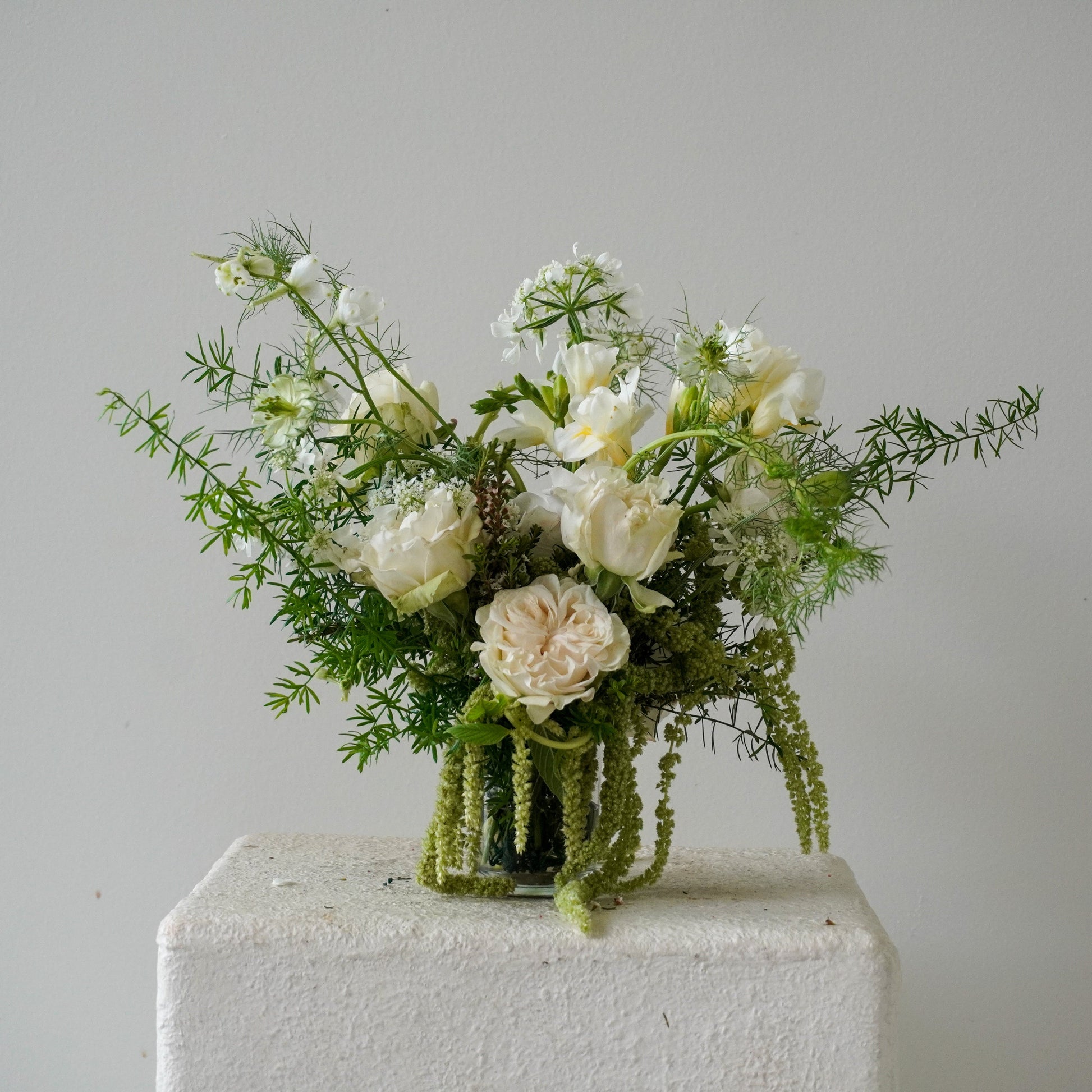 Bouquet of white flowers and greenery in a clear vase on a white pedestal by goodroots.la